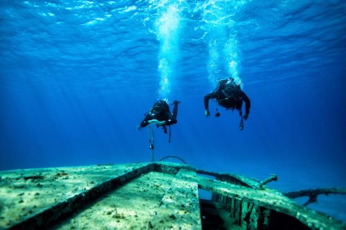 Two scuba divers exploring huge ship wreck on the sand sea bottom.